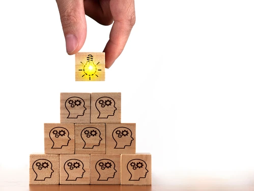 Hand dropping block with light bulb on stack of blocks with gears turning in human heads